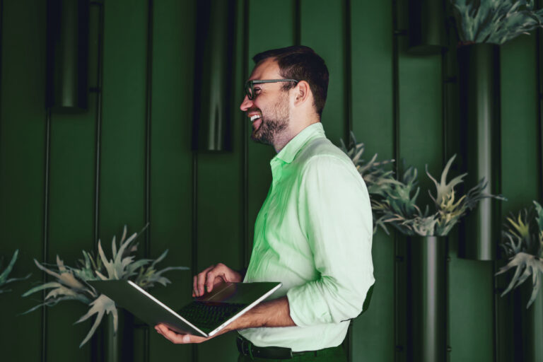 Homem branco em pé e com um notebook aberto em sua mão. Ele está sorrindo e há plantas decorando a parede.