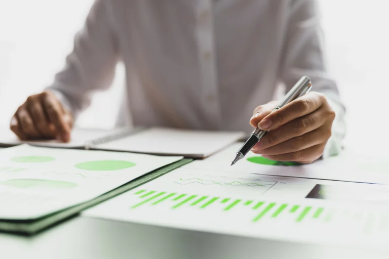 Pessoa fazendo cálculo de cota capital em uma mesa com um computador, alguns papéis com gráficos financeiros e cadernetas.