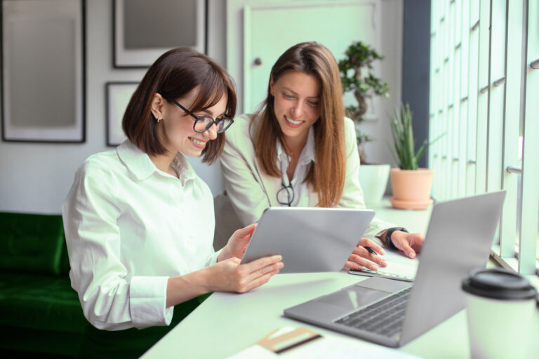 Duas mulheres brancas conversando em um escritório. Uma delas usa um tablet. Há um notebook na mesa.