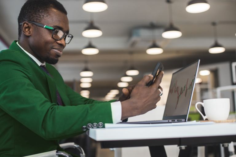 Homem usando o celular com os braços apoiados na mesa onde há um notebook aberto com gráficos financeiros em sua tela.