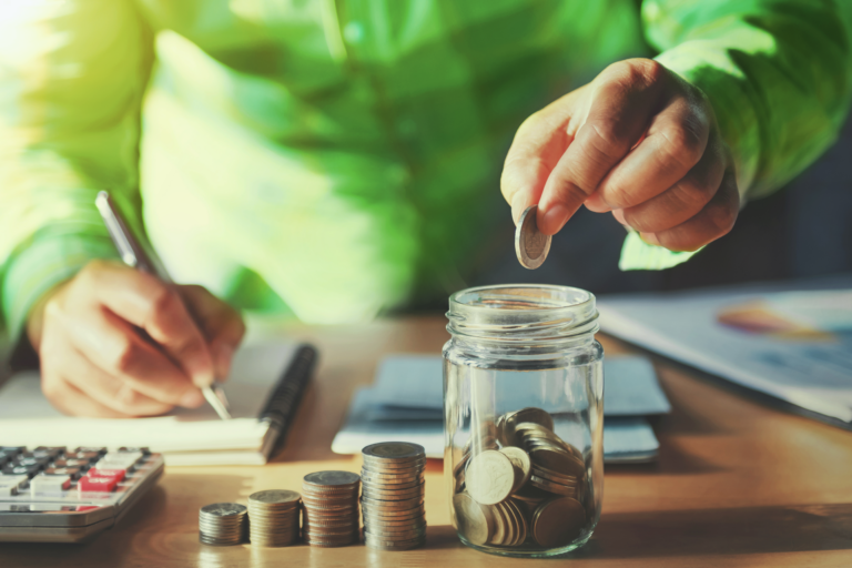 Pessoa depositando moedas num pote sobre uma mesa. Ela faz anotações em um caderno. Há uma calculadora ao lado.