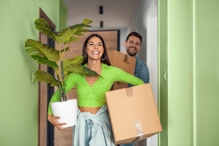 Casal chegando em sua casa própria. A mulher, à frente, carrega uma caixa e um vaso com plantas, o homem, atrás, com uma caixa.