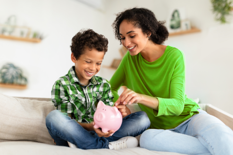 Criança colocando moeda em seu cofrinho junto com a mãe, representando a aprendizagem sobre educação financeira desde a infância.