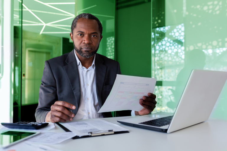 Homem negro, investidor, em um escritório. Ele está segurando um papel. Há um notebook e uma calculadora ao lado dele com investimento em royalties