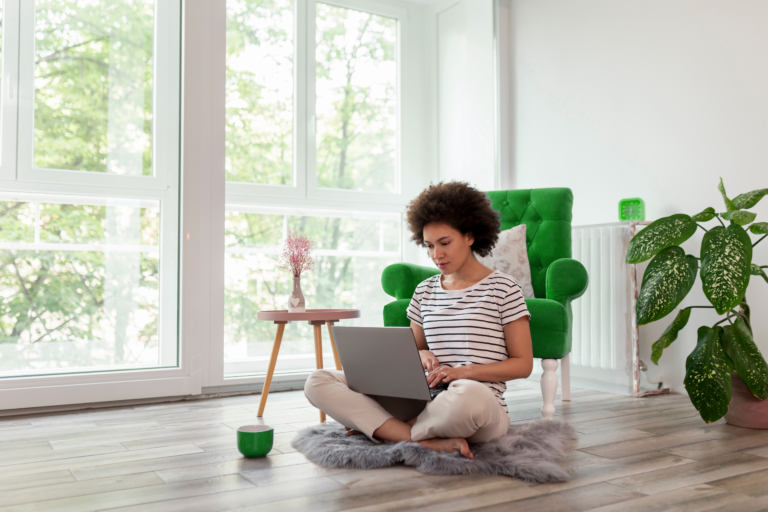 Mulher negra sentada no chão da sala. Ela está usando um notebook. Há plantas ao lado dela e uma xícara com café. Ela tem um negócio e está buscando opções de investimento para autônomo.