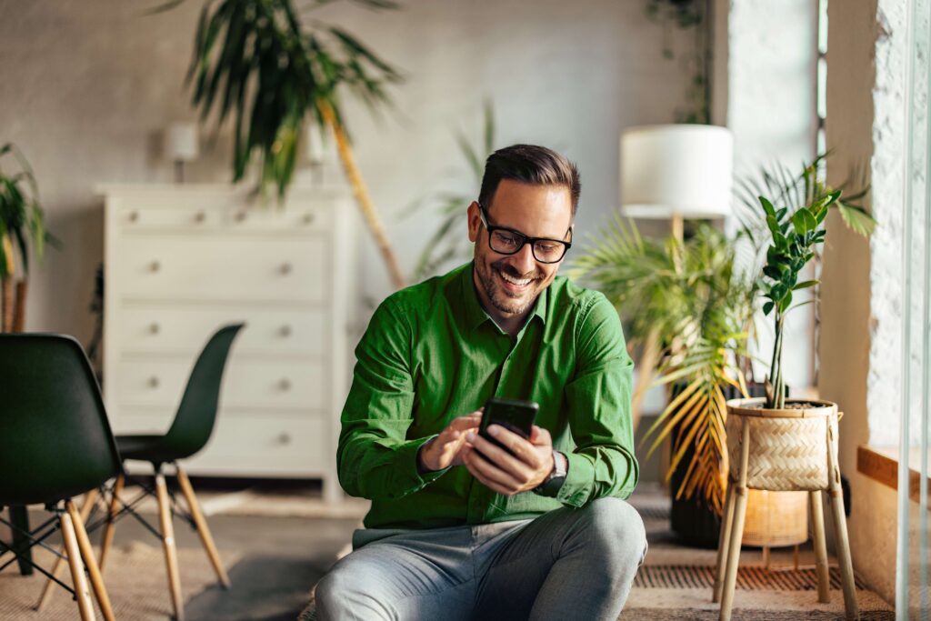 homem de camisa verde sentado em escritório sorrindo enquanto olha para tela de celular após fazer um pix em conta pj