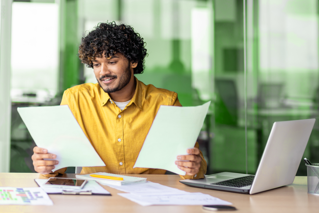 Homem de cabelos cacheados em uma mesa de escritório buscando informações sobre investimentos para PJ. Ele está segurando papéis e há um notebook ao lado dele.