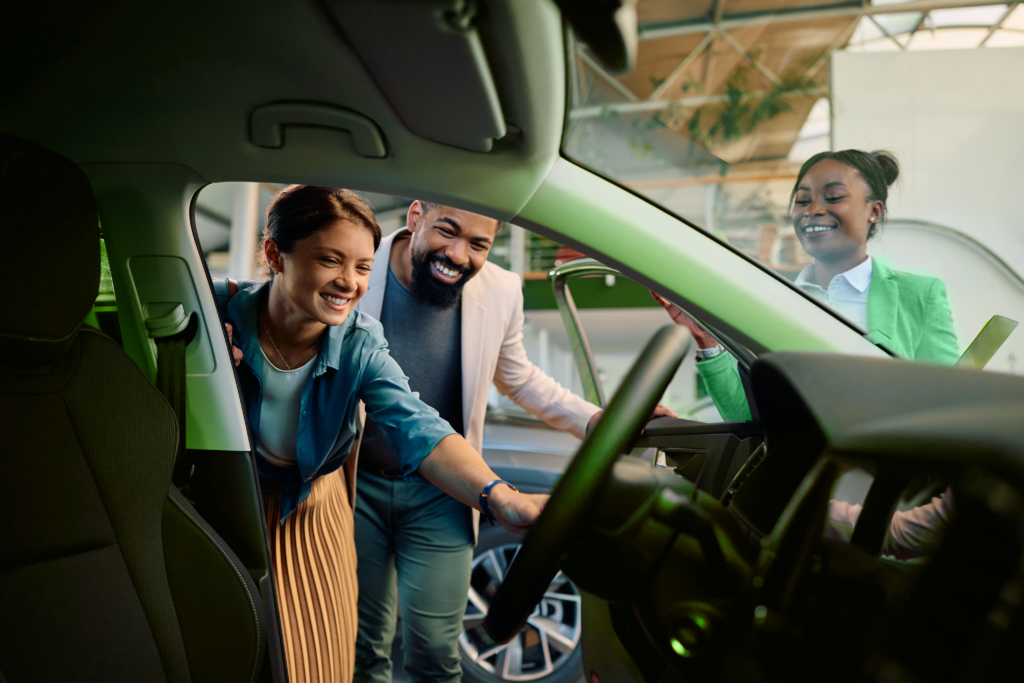 um casal sorrindo olhando para dentro de um carro com uma profissional próxima a eles sorrindo também referente a refinanciar veículo.