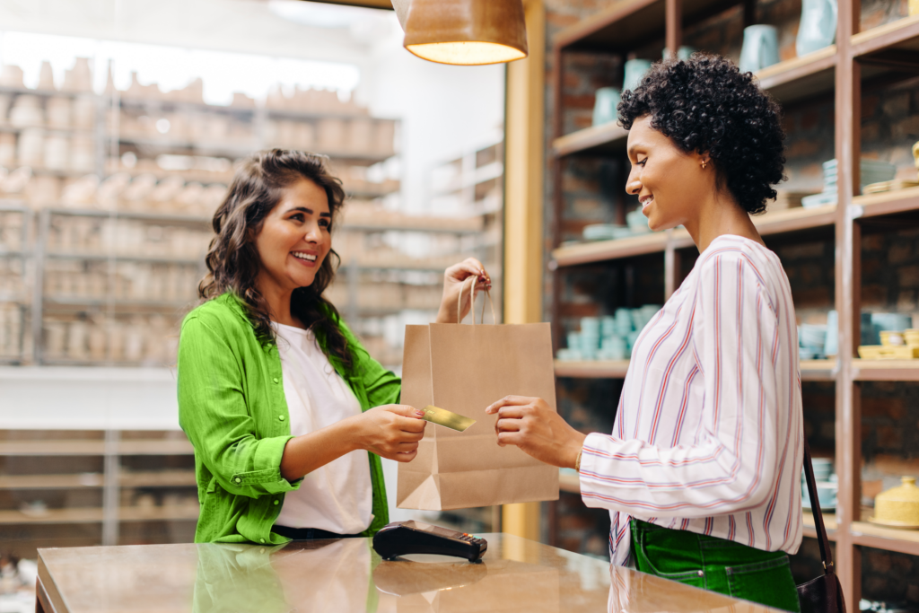 dona de loja realizando venda entregando compra e seu cartão de crédito para cliente representando o consumo consciente.