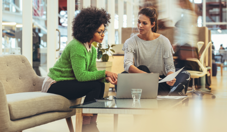 Duas mulheres conversando, sentadas, analisando gráficos financeiros em um notebook. Elas estão em um espaço público.