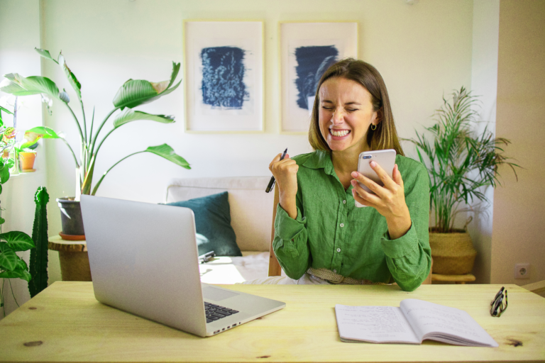 Mulher com celular nas mãos e laptop na frente, sentado em uma mesa feliz, comemorando.