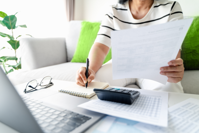 Mulher calculando a tabela IOF em sua casa. Ela está com papeis financeiros, uma calculadora e um notebook.