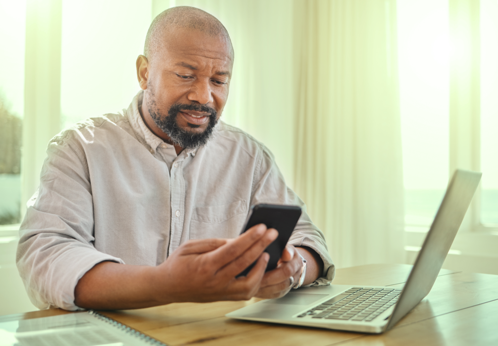 Homem negro com celular nas mãos e laptop na frente, sentado em uma mesa