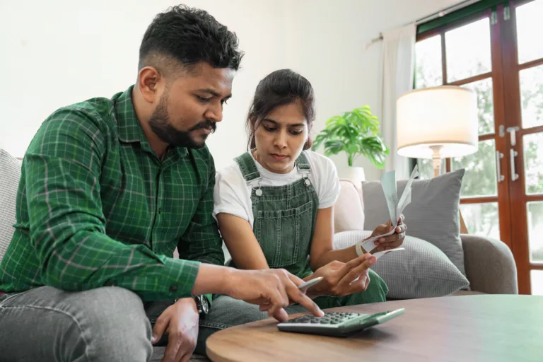 Casal no sofá fazendo contas na calculadora e segurando papéis ajustando detalhes para uma melhor economia doméstica