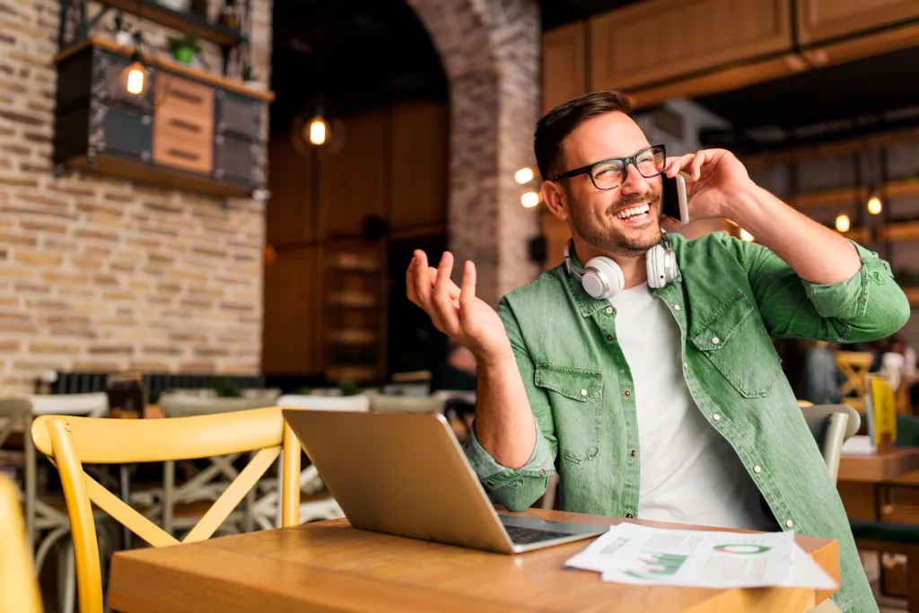 profissional em trabalho como freelancer falando ao telefone sorrindo enquanto está sentado na mesa de um café com um notebook na mesa